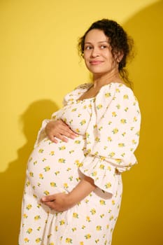Attractive middle-aged African American gravid woman, gently holding her belly, smiling looking aside, isolated yellow background. Pregnancy. Maternity. Childbearing. Expecting a baby. Motherhood
