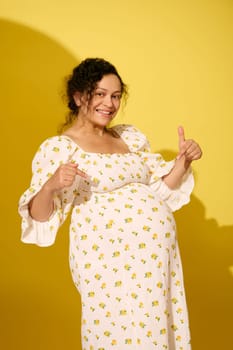 Beautiful pregnant woman dressed in summer sundress, points fingers at her belly, smiles looking at camera, isolated over yellow studio background. Happy carefree pregnancy and maternity concept