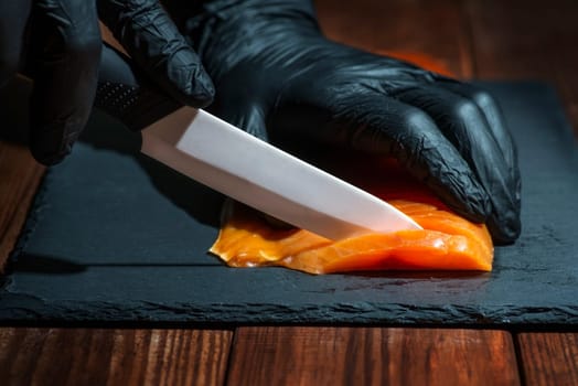 Chef use knife to cutting salmon fillet on black board at professional kitchen. Closeup chef hands in black gloves slicing red fish slice.