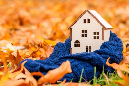 Toy wooden house is wrapped in a warm scarf on the autumn leaves background. Warm house concept. Preparing the house for the cold.