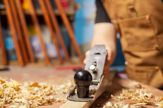 Carpenter in an apron planing a wooden board with a planer, workplace.