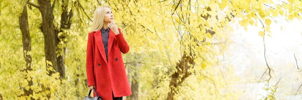 A young blonde woman walks around the autumn city in a red coat. The concept of urban style and lifestyle.