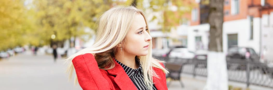 A young blonde woman walks around the autumn city in a red coat. The concept of urban style and lifestyle.