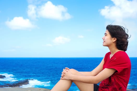 Young woman sitting on large rock cliff  by Hawaiian  ocean , looking out over horizon
