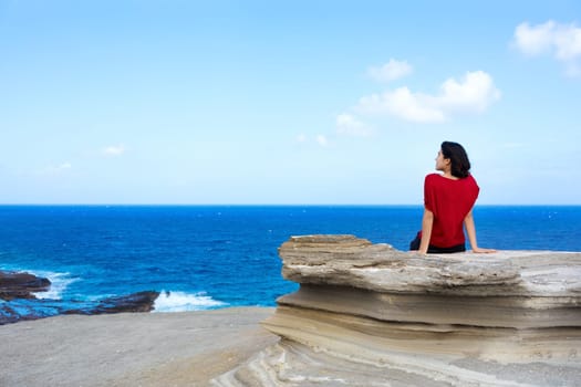 Young woman sitting on large rock cliff  by Hawaiian  ocean , looking out over horizon