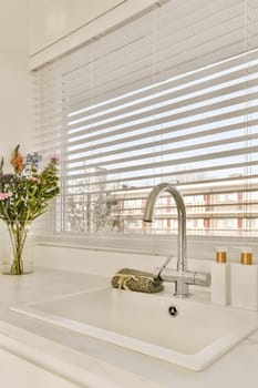 some flowers in a vase on the kitchen sink with white blinds behind it and an open window that looks out to the city