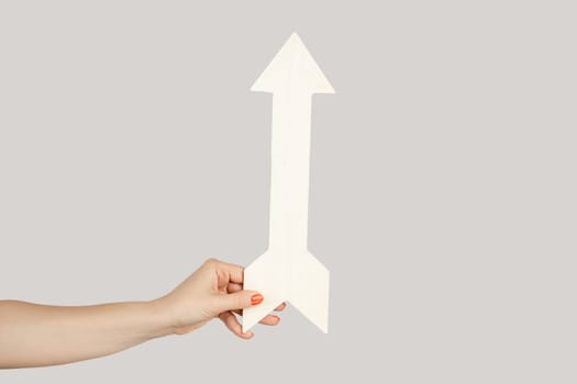 Closeup of woman hand holding white arrow indicating direction upwards, symbol of increase. Indoor studio shot isolated on gray background.