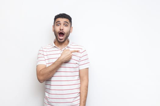 Portrait of astonished shocked bearded man wearing striped t-shirt standing pointing aside at copy space for promotion, advertisement area. Indoor studio shot isolated on gray background.