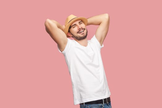 Portrait of satisfied relaxed handsome bearded hipster man in white T- shirt and casual hat standing with raised hands, looking at camera. Indoor studio shot isolated on pink background.