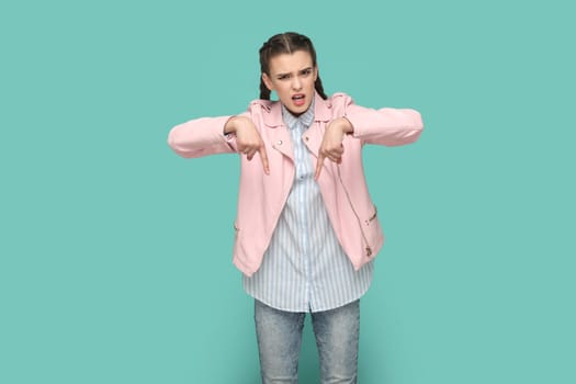 Portrait of strict bossy teenager girl with braids wearing pink jacket pointing both index fingers down, saying here and right now. Indoor studio shot isolated on green background.