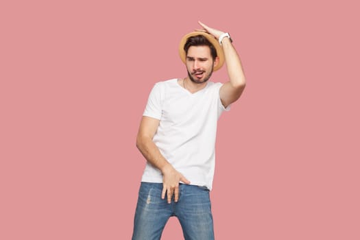 Portrait of excited playful handsome bearded man in white T-shirt and hat dancing, having funny pose, spending time on party. Indoor studio shot isolated on pink background.