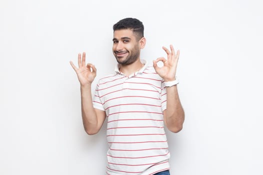 Everything ok. Portrait of attractive positive smiling bearded man wearing striped t-shirt standing showing okay gesture, agreed. Indoor studio shot isolated on gray background.