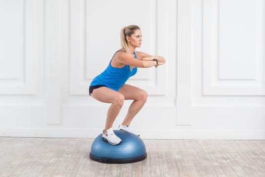 Side view portrait of sporty attractive woman wearing black shorts and blue top working in gym doing exercise in bosu balance trainer, squats on fitness ball, holding balance. Indoor studio shot.