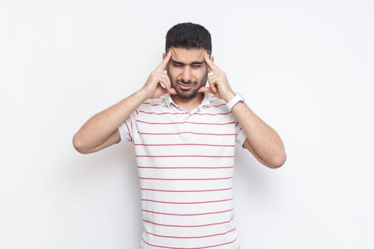 Portrait of sick unhealthy ill bearded man wearing striped t-shirt standing suffering headache, massaging his temples, frowning face. Indoor studio shot isolated on gray background.