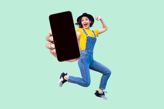 Portrait of happy surprised hipster girl in overalls, T-shirt and hat jumping and looking at camera, showing big empty smart phone display. Indoor studio shot isolated on light green background.