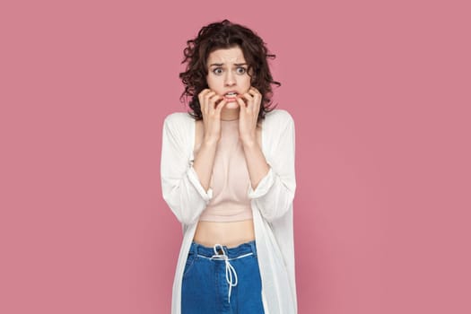 Portrait of nervous worried young adult woman with curly hair wearing casual style outfit, having troubles, biting her fingernails. Indoor studio shot isolated on pink background.