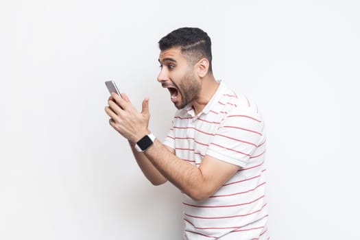 Side view portrait of amazed excited handsome bearded man wearing striped t-shirt standing using cell phone, reading shocking news. Indoor studio shot isolated on gray background.