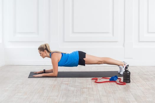 Side view of sporty strong young blonde woman wearing black shorts and blue top standing on perfect plank position, training full body and abs, training in gym. Indoor studio shot.