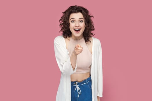 Portrait of amazed surprised woman with curly hairstyle wearing casual style outfit choosing you, looking at camera with big eyes and pleasant surprise. Indoor studio shot isolated on pink background.