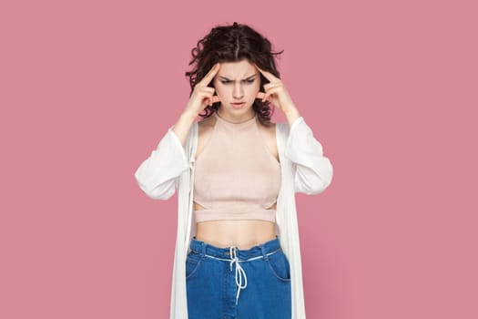 Portrait of confused puzzled woman with curly hair wearing casual style outfit thinking, keeps hands on temples, having serious expression. Indoor studio shot isolated on pink background.