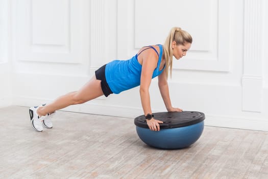 Full length portrait of concentrated woman in black shorts and blue top working in gym doing plank for abdominal muscles on bosu balance trainer, holding balance on fitness ball. Indoor studio shot.
