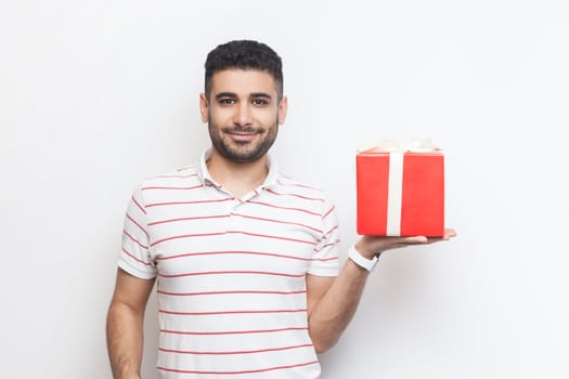 Portrait of delighted cheerful handsome bearded man wearing striped t-shirt standing showing red wrapped present box, gift for birthday. Indoor studio shot isolated on gray background.