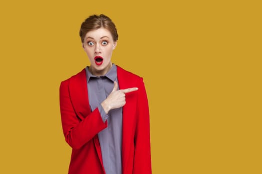 Portrait of amazed astonished woman with red lips standing pointing finger aside at copy space for advertisement, wearing red jacket. Indoor studio shot isolated on yellow background.