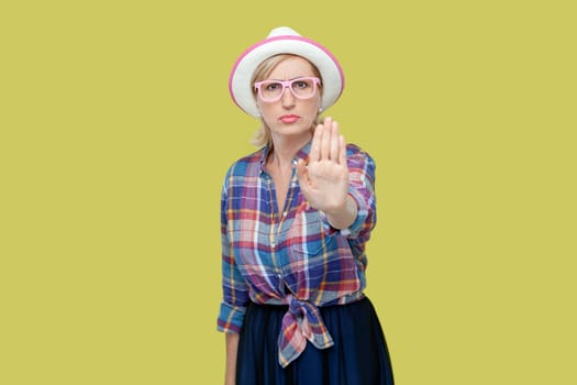 Portrait of serious senior woman wearing checkered shirt, hat and eyeglasses raises palm in stop or prohibition gesture, says no wait sec, hold on. Indoor studio shot isolated on yellow background