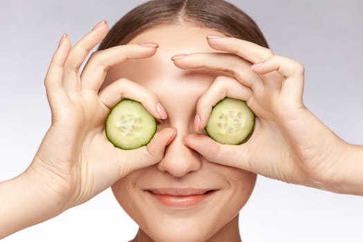 Closeup portrait of joyful beautiful woman smiling hiding eyes behind cucumber slices beauty treatment skin care. Indoor studio shot isolated over gray background.