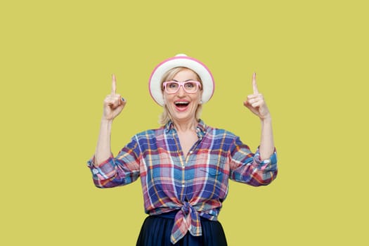 Senior woman wearing checkered shirt, hat and eyeglasses looks with widely opened mouth up, sees something amazed, indicates with fore fingers. Indoor studio shot isolated on yellow background.