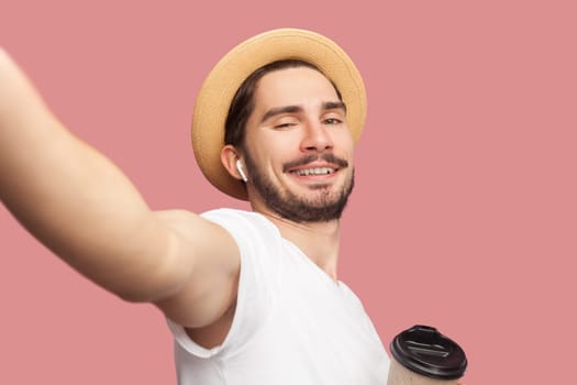 Portrait of handsome cheerful joyful bearded man in white T-shirt and hat standing drinking coffee, looking smiling at camera, POV. Indoor studio shot isolated on pink background.