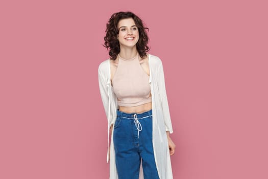 Portrait of extremely happy delighted smiling woman with curly hair wearing casual style outfit standing looking away with positive expression. Indoor studio shot isolated on pink background.