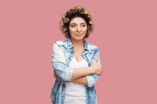 Portrait of positive pretty young adult woman with curly hairstyle wearing blue shirt standing with folded hands, looking away and dreaming. Indoor studio shot isolated on pink background.