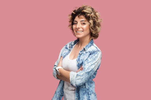 Portrait of confident happy smiling woman with curly hairstyle wearing blue shirt standing with crossed arms, looking at camera with positive expression. Indoor studio shot isolated on pink background
