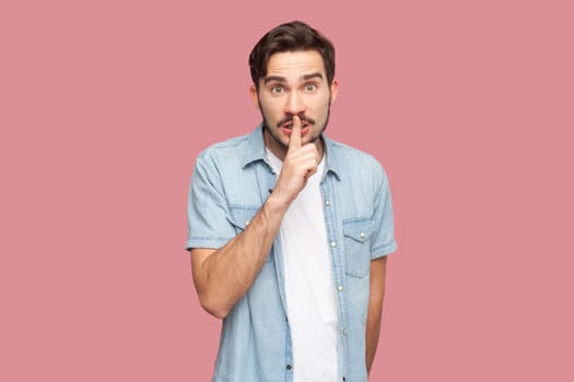 Portrait of stupefied secret bearded man in blue casual style shirt standing makes gesture quietly, asks remain silent, gossips about something. Indoor studio shot isolated on pink background.