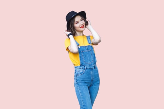 Portrait of cute charming hipster woman in blue denim overalls, yellow T-shirt and black hat, looking at camera with positive expression. Indoor studio shot isolated on light pink background.