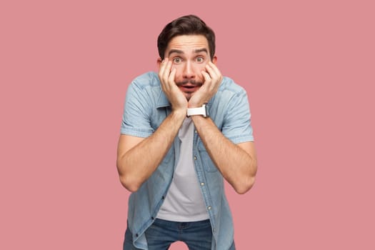 Portrait of shocked astonished bearded man in blue casual style shirt standing with hands on cheeks, looking at camera with big eyes. Indoor studio shot isolated on pink background.