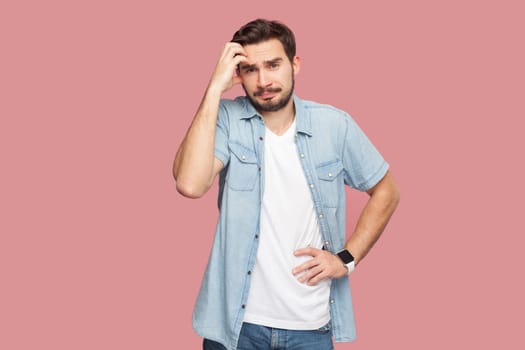 Portrait of puzzled bearded man in blue casual style shirt standing looks confusingly at camera, keeps hand on hip, rubbing his head. Indoor studio shot isolated on pink background.