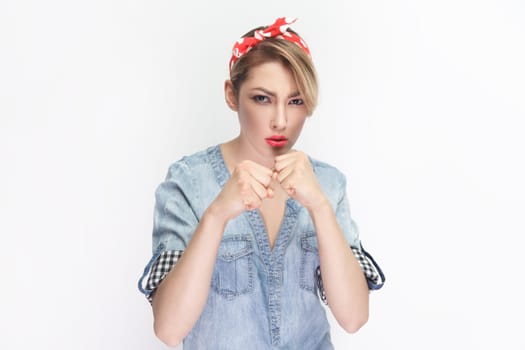 Portrait of angry irritated blonde woman wearing blue denim shirt and red headband standing with clenched fists, fighting with somebody. Indoor studio shot isolated on gray background.
