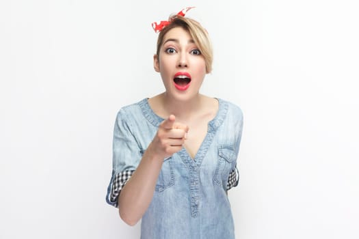 Portrait of surprised shocked blonde woman wearing blue denim shirt and red headband standing, pointing at you, looking with big eyes. Indoor studio shot isolated on gray background.