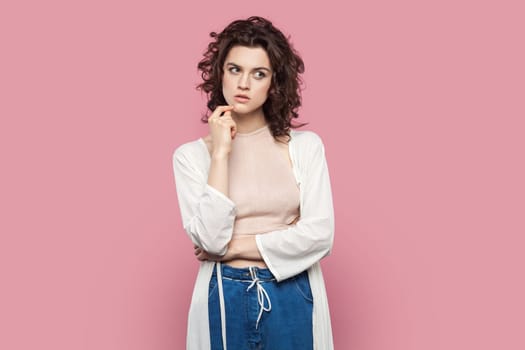 Portrait of pensive attractive woman with curly hairstyle wearing casual style outfit, holding chin, looking away, thinking about her job. Indoor studio shot isolated on pink background.