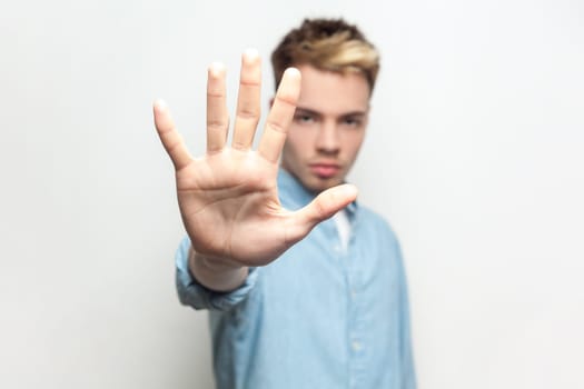 Portrait of serious bossy confident attractive man wearing denim shirt standing with outstreched hand, showing stop gesture with palm. Indoor studio shot isolated on gray background.