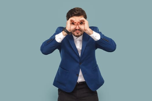 Portrait of attractive man with mustache standing with binocular gesture, looking far away, expecting good future, wearing official style suit. Indoor studio shot isolated on light blue background.