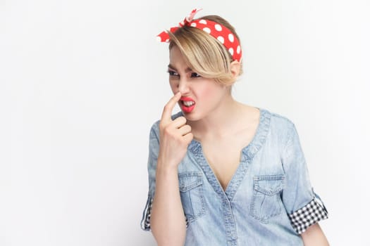 You are liar. Portrait of funny foolish blonde woman wearing blue denim shirt and red headband standing touching her nose, frowning face. Indoor studio shot isolated on gray background.