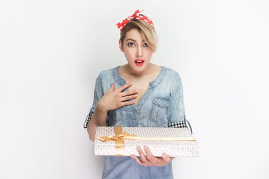 Portrait of surprised blonde woman wearing blue denim shirt and red headband standing holding present box, gets unexpected gift. Indoor studio shot isolated on gray background.