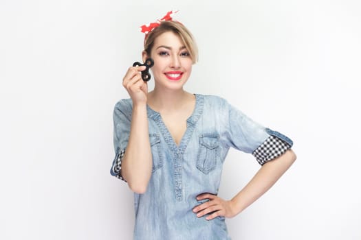 Portrait of smiling cheerful joyful blonde woman wearing blue denim shirt and red headband standing holding spinner, keeps hand on hips. Indoor studio shot isolated on gray background.