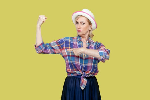 Portrait of mature woman wearing checkered shirt and hat having serious bossy expression, raises arms and showing pointing at her muscles. Indoor studio shot isolated on yellow background.