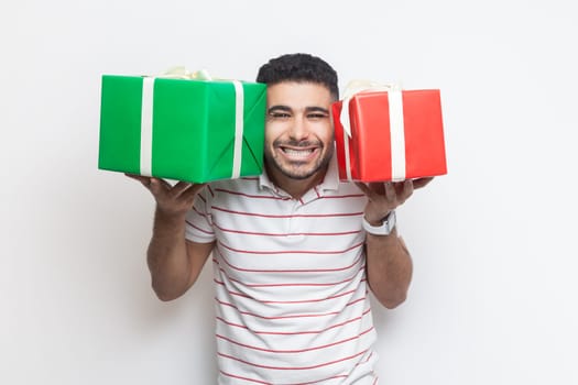 Portrait of extremely happy smiling bearded man wearing striped t-shirt standing with two present boxes, looking at camera with toothy smile. Indoor studio shot isolated on gray background.
