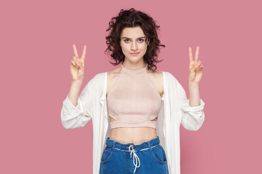 Portrait of attractive beautiful young adult woman with curly hair wearing casual style outfit standing looking at camera, showing v sign. Indoor studio shot isolated on pink background.