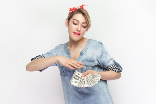 Portrait of rich satisfied blonde attractive woman wearing blue denim shirt and red headband standing throwing money, having big salary. Indoor studio shot isolated on gray background.